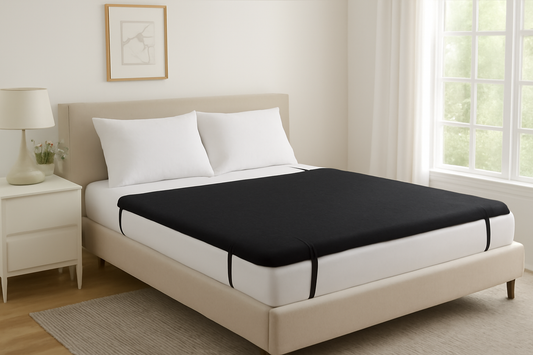 A simple bed with a black fitted sheet, white pillows, and a lamp beside it in a bright room.