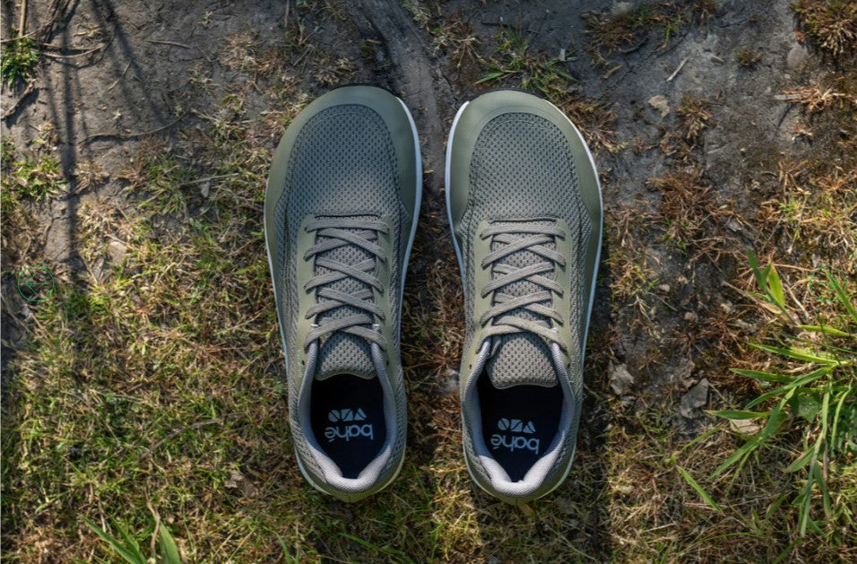 A pair of Bahe grounding shoes in an earthy green color placed on a natural outdoor surface with grass, dirt, and tree roots, emphasizing their connection to nature and grounding benefits