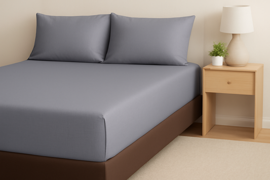 A neatly made bed with gray sheets and brown base, complemented by a nightstand with a lamp.